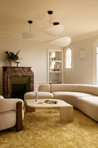Elegant living room featuring a large curved bouclé sofa, marble fireplace with green tile interior, travertine coffee table, and yellow shaggy carpet under modern sculptural pendant lights.