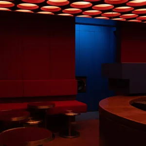 Interior of a modern nightclub lounge with red velvet banquette seating, round metallic tables, and a ceiling covered in glowing red circular light fixtures against a deep blue wall.