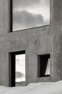 Close-up view of a raw concrete building facade featuring large and small black-framed windows, with snow piled at the base and a snowy mountain visible in the distance.
