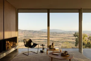 Modern living room interior featuring a black leather sling chair, round oak coffee tables, built-in wood storage, and floor-to-ceiling windows overlooking a vast mountain and valley landscape.