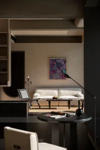 Modern minimalist living room featuring textured beige plaster walls, a white sofa, black woven chair, and a large colorful abstract painting.