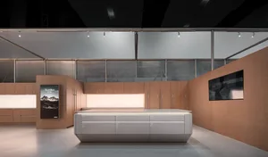 Modern minimalist kitchen featuring a large grey central island, extensive light wood cabinetry, under-cabinet lighting, and a digital art display in a spacious, high-ceilinged room.