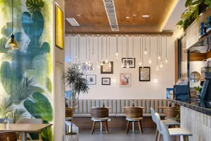 Modern cafe interior with large tropical leaf mural on the left, multiple wooden pendant lights hanging from a wooden ceiling, upholstered banquette seating, and round wooden tables with white chairs.