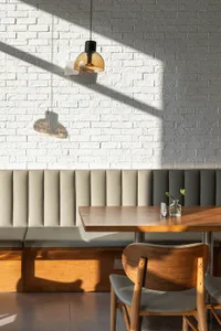 Modern restaurant interior featuring a white painted brick wall, beige upholstered booth seating, solid oak dining table, wooden chair with grey leather seat, and a glass pendant lamp casting strong sunlight shadows.