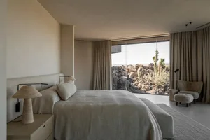Modern minimalist bedroom with neutral linen bedding, travertine table lamp, and large glass windows overlooking a rocky desert landscape with cacti.