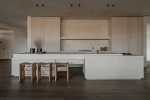 Modern minimalist kitchen featuring a large concrete island with integrated sink and cooktop, three curved wooden bar stools, light wood cabinetry, and dark wood flooring.