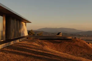 Modern minimalist concrete terrace extending from a contemporary house, featuring a single black chair overlooking golden-hour mountain landscape