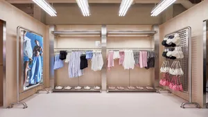 Modern minimalist clothing boutique interior with light plywood walls, stainless steel clothing racks, shoe display shelves, hat and bag grid wall, and a large fashion photograph on a rolling metal frame under bright fluorescent lighting.