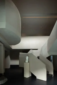 Modern sculptural concrete staircase with integrated lighting, featuring a white marble bust on a stone pedestal against a dark floor in a minimalist interior.