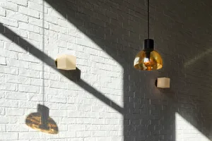 White painted brick wall illuminated by dramatic diagonal sunlight, featuring a large amber glass pendant lamp and two small cube-shaped wall sconces casting strong geometric shadows.