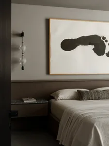 Modern minimalist bedroom featuring a walnut wood panelled headboard, neutral linen bedding, marble nightstand, modern glass wall sconce, and large abstract black ink painting.
