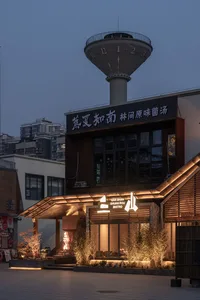 Night view of San Shan Shuan Rou Bistro with illuminated corten steel facade, wooden slats, and lush landscaping under a large concrete water tower.