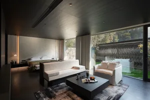 Modern minimalist bedroom sitting area featuring a white sofa, white armchair, dark wood coffee table, and large floor-to-ceiling windows overlooking a private garden with a patterned fence.