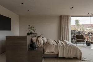 Modern minimalist bedroom featuring a low wooden platform bed with beige linen bedding, textured wood headboard, and large glass doors opening to a rocky desert landscape with yucca plants.
