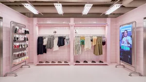 Interior view of a modern clothing boutique with bright pink tiled walls, metal clothing racks, shoe displays, and a large digital screen on the right.