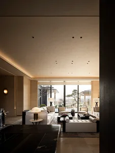 Modern luxury living room featuring travertine walls, large white sectional sofa, dark marble coffee tables, and floor-to-ceiling windows overlooking a Japanese garden with bonsai trees.