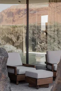 Two modern walnut wood armchairs with light grey cushions and matching ottomans placed in a minimalist living space with floor-to-ceiling glass walls overlooking a rugged desert mountain landscape with volcanic rock formations.