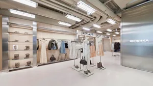 Interior view of a Bershka clothing store featuring white mannequins, clothing racks, wooden shelving with boots, exposed ceiling ducts, and a large stainless steel wall with the brand logo.