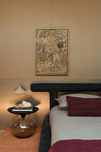 Modern bedroom corner with dark boucle upholstered headboard, terracotta knit throw, black side table with glass base, and framed antique tapestry with jewel embellishments on a neutral wall.