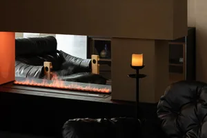 Modern living room featuring a long linear electric fireplace, black leather sofas, wooden speakers, and a lit candle on a black stand.