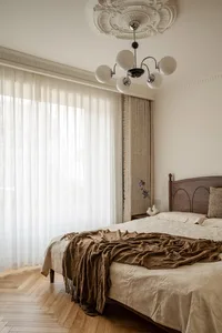 A tranquil bedroom featuring a dark walnut wooden bed, ornate white ceiling medallion with modern chandelier, sheer white curtains, textured beige drapes, and a bed layered with cream patterned linens and a rumpled brown throw blanket on herringbone wood flooring.