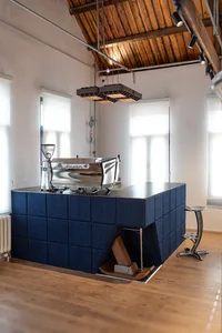 Modern industrial-style coffee station featuring a large chrome espresso machine on a blue fabric-paneled counter with wooden floor and exposed wooden ceiling beams.