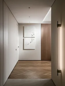 Modern minimalist hallway featuring light oak herringbone parquet flooring, two large textured white abstract artworks on a white wall, vertical wall sconce, and warm wood door under recessed ceiling lighting.