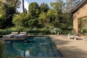 Modern outdoor terrace with a dark infinity swimming pool, concrete deck, outdoor lounge beds, wireframe chairs, and a dense green forest garden in the background.