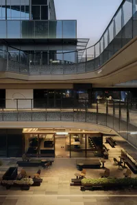 Modern multi-story commercial building interior with curved glass facade, outdoor plaza seating, and a ground-floor Oasis Coffee shop at dusk.