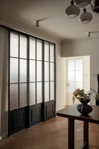 Modern dining area featuring a large black steel-framed textured glass partition wall, dark wood dining table with black vase and dried flowers, and warm herringbone oak flooring.