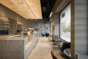 Modern minimalist cafe interior with grey concrete walls, tiled service counter, wooden ceiling panels, grey upholstered lounge chairs, and large windows overlooking greenery.