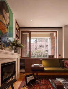 Modern eclectic living room with a travertine fireplace on the left, a large colorful portrait painting, built-in wooden bookshelves, an olive green velvet sofa, and a large window overlooking blooming cherry blossom trees against a brick building.