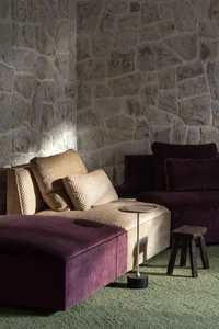A modern lounge setting featuring a plum velvet chaise and ottoman paired with a beige checkered sofa, set against a textured stone wall with dramatic uplighting, accompanied by a minimalist side table and rustic wooden stool on green carpet.