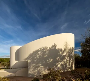 Modern curved white plaster building with dramatic tree shadows cast by Mediterranean sunlight against a deep blue sky.