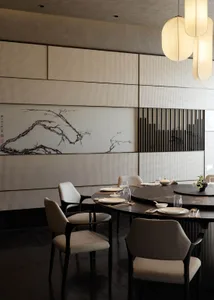 Modern Chinese restaurant interior with a large round dark marble dining table, cream upholstered walnut chairs, large traditional Chinese ink painting of a plum blossom branch on a textured wall, and hanging spherical pendant lights.