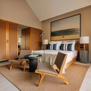 Modern coastal bedroom with light wood teak furniture, woven rattan lounge chairs, neutral linen bedding with blue striped pillows, large beach landscape artwork, and a textured jute rug on tiled floor.