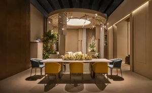 Modern dining room featuring a long rectangular table surrounded by yellow and blue patterned velvet chairs, a large dried floral centerpiece, and a dramatic circular ceiling installation with branching light fixtures.