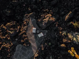 Aerial view of two white lounge chairs on a dark basalt stone platform surrounded by black volcanic rocks and desert plants under warm sunlight.