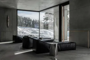 Modern minimalist living room with raw concrete walls, large floor-to-ceiling windows overlooking a snowy mountain forest, black leather modular sofas, and brushed metal side tables.