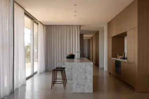 Modern minimalist kitchen with a large grey terrazzo island, wooden bar stools, floor-to-ceiling sheer curtains, and warm oak cabinetry along the right wall.
