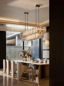 Modern luxury dining room featuring a large rectangular alabaster and brass chandelier, a marble dining table, four textured boucle dining chairs, and a large floral arrangement with a view to an outdoor courtyard.