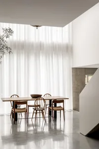 Modern minimalist dining room featuring a light oak rectangular table surrounded by six wooden spindle-back chairs with woven seats, set against a backdrop of floor-to-ceiling sheer white curtains, polished concrete floor, and a raw concrete staircase on the right.