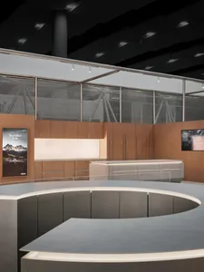 Modern luxury boutique interior featuring a large curved grey reception island in the foreground, warm oak cabinetry and backlit display wall in the background, with large windows covered by horizontal blinds.