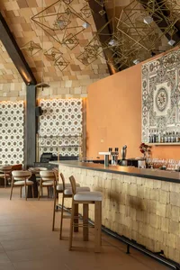 Modern restaurant bar featuring a long counter clad in light wood shingles, wooden bar stools, terracotta accent wall, patterned tile art, and numerous geometric brass pendant lights hanging from a wood-paneled ceiling.