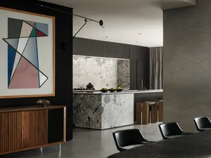 Modern dark kitchen interior with large white and grey terrazzo island, dark wood cabinetry, black dining chairs, and a large colorful geometric abstract painting on a black wall.