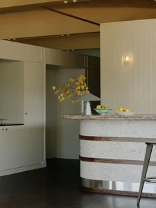 Modern kitchen with a curved travertine island featuring horizontal oak bands, a grey table lamp, fruit bowls, and a wall sconce against a white tiled wall.