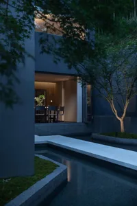 Night view of a modern concrete house courtyard with a linear water feature, moss garden, small tree, and warmly lit indoor dining area visible through large openings.