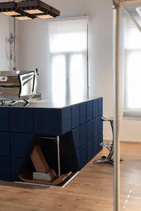 Modern coffee bar with navy blue velvet upholstered base, stainless steel espresso machine, chrome bar stool, and large geometric pendant light hanging above a light wood floor.