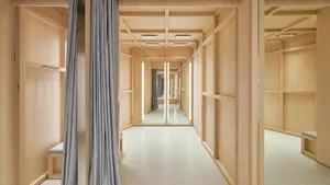 Modern minimalist walk-in dressing room lined with light oak cabinetry, grey curtains, built-in seating, and multiple vertical mirrors creating an infinite hallway effect.