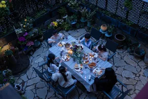 Overhead view of a family of six enjoying dinner at a long table set on a rustic stone-paved courtyard at dusk, surrounded by numerous potted flowers and plants.